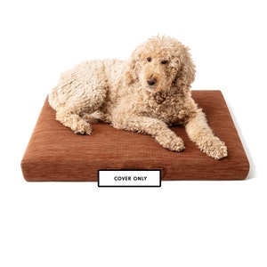 May include: A light brown dog bed with a brown and white striped pattern. The dog bed has a label that says "COVER ONLY". A light brown dog is lying on the bed.