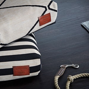 May include: Two dog beds stacked on top of each other. The top bed is beige with a black geometric pattern and the bottom bed is beige with black stripes. Both beds have a brown leather patch with the text "ALTUZARRA x LAYR" on them.