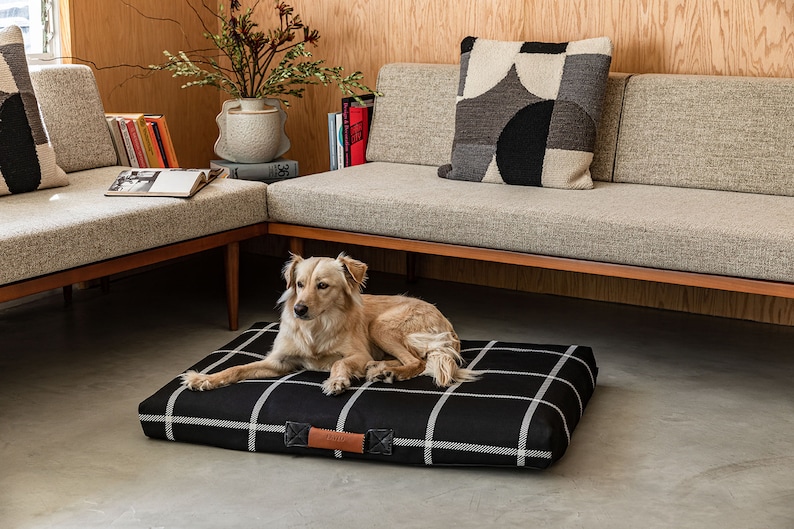 May include: A dog bed with a black and white checkered pattern. The bed has a rectangular shape with a brown leather handle. A golden retriever dog is lying on the bed. The bed is in a living room with a sofa and pillows.