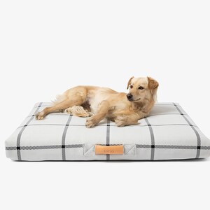 May include: A light-colored dog rests on a square dog bed. The bed has a white background with a black plaid pattern. A leather tag with the word "Lotto" is attached to the bed.