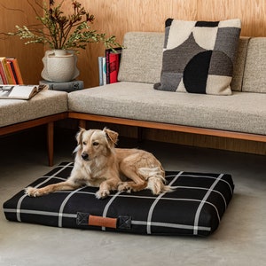 May include: A black and white plaid dog bed with a brown leather handle. A golden dog is lying on the bed in a living room setting.