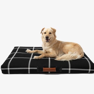 May include: A golden-colored dog rests on a black and white plaid dog bed. The bed has a rectangular shape with a brown leather handle on one side. The dog is looking at the camera.