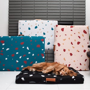 May include: Three dog beds with a terrazzo pattern in blue, pink, and white. A dog is lying on the black dog bed with white spots.