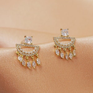 Może przedstawiać: Para złotych kolczyków z designem w kształcie łzy i przezroczystymi kamieniami cyrkonowymi. Kolczyki mają zwisający design z wieloma kamieniami.