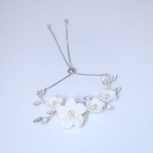 Peut inclure: Un bracelet floral blanc avec une chaîne argentée et un fermoir réglable. Le bracelet présente de délicates fleurs blanches avec des accents de perles et des feuilles bleues irisées.