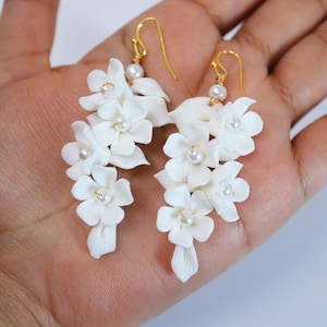 Peut inclure: Une paire de boucles d'oreilles pendantes en forme de fleurs blanches avec des crochets dorés et des accents de perles. Les boucles d'oreilles sont en pâte polymère et ont la forme de fleurs délicates.