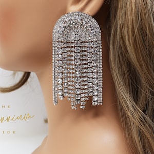 Może przedstawiać: Para dużych, srebrnych kolczyków wiszących ozdobionych kryształami. Kolczyki mają kształt łzy z dużym, okrągłym kryształem na górze i kaskadą mniejszych kryształów zwisających w dół.