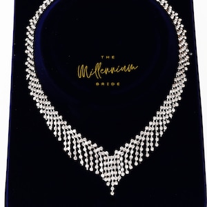 Peut inclure: Un collier argenté avec un design en cascade de petits strass transparents. Le collier est présenté sur un fond de velours bleu foncé. Le texte "THE Millennium BRIDE" est visible en lettres dorées.