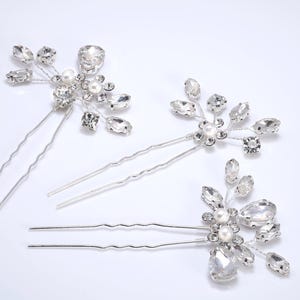 Puede incluir: Tres horquillas plateadas con detalles de cristal y perlas. Cada horquilla presenta un diseño floral con cristales claros y facetados y una perla central. Ideales para peinados de novia o formales.
