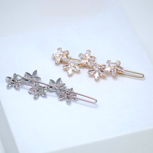 May include: Two floral hair clips. One is gold-toned, the other is silver-toned. Each clip features a row of three flower-shaped embellishments with clear, sparkling crystals. The clips are designed to be worn in the hair.