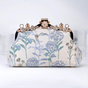 May include: An off-white clutch with a floral pattern in shades of blue and green. The clasp and trim are gold-toned, with sparkling crystal accents. The bag has a structured shape and a delicate, feminine design.