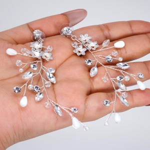 Peut inclure: Une paire de boucles d'oreilles argentées d'inspiration florale. Chaque boucle d'oreille présente un groupe de petits cristaux transparents et des accents de fleurs blanches. Des perles claires en forme de goutte et des cristaux étincelants sont disposés le long de tiges délicates et ramifiées.