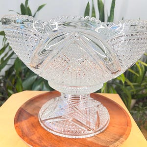 Könnte beinhalten: Eine transparente, geschliffene Bowleschüssel mit Sockel, auf einem runden Holztablett. Die Schüssel weist komplizierte geometrische Muster und einen gewellten Rand auf. Die Schüssel ist zum Servieren von Getränken bestimmt.