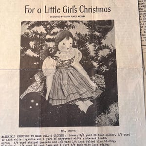 May include: A vintage black and white printed pattern sheet titled For a Little Girl's Christmas, designed by Edith Flack Ackley. It features an illustration of a cloth doll in a dress and apron, with detailed material requirements listed below.