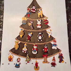 Op de afbeelding: Een donkergroene kerstboom-wandhanger van vilt, versierd met gouden franjes en diverse kleine, kleurrijke vilten ornamenten, waaronder een kerstman, een konijn, een panda, een sneeuwpop, een uil, een kerkje en een clown.