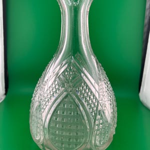 May include: A clear glass decanter with a wide, patterned base and a slender neck. The decanter features a detailed, cut-glass design with diamond and square patterns. The clear glass allows light to pass through, highlighting the intricate details.