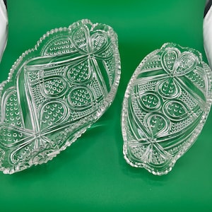 May include: Two clear, pressed glass serving dishes with a clover-like pattern. The dishes have a scalloped edge and are set against a green background. The larger dish is approximately 10 inches long, and the smaller dish is about 7 inches long.