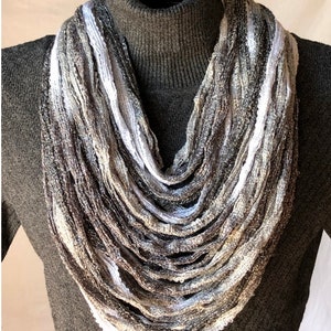 May include: A long, gray, white, and silver infinity scarf made of thin, woven fabric. The scarf is draped around a turtleneck sweater.