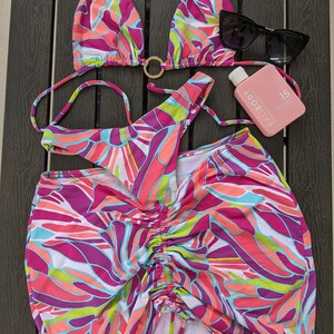 May include: A pink, green, and purple patterned bikini top, bottom, and skirt. The skirt has a drawstring waist. The bikini top has a gold ring detail. A pink bottle of sunscreen with the text "Bali Body" and "SPF 15" is in the image.