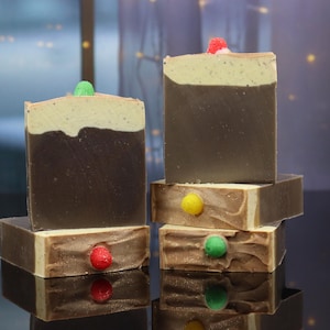 May include: Stacked handmade soaps with a dark brown base and a light tan top. Each soap is topped with a colorful decorative embellishment. The soaps are arranged on a reflective surface, creating a visually appealing display.