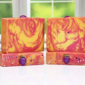 May include: Two bars of handmade soap. The top bars have a swirl pattern of yellow, orange, and pink. The bottom bars are pink with purple strawberry embellishments and glitter. The background is a window.