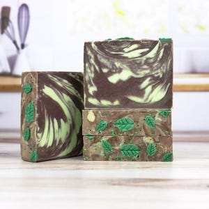 May include: Stacked bars of handmade soap with a brown and green swirl pattern. The bottom bars have green leaf embellishments. The soaps are displayed on a light-colored surface with a kitchen background.