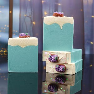 May include: Several bars of handmade soap are stacked, featuring a two-tone design of light blue and cream. Each bar is topped with a small, decorative cluster of purple grapes. The soaps are arranged on a reflective surface.