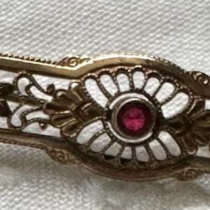 May include: A gold-coloured bar pin with a central red gemstone and floral filigree design. The pin has a white enamel background and is set against a white, textured surface. The pin is approximately 5 cm long.