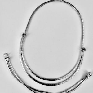 May include: A silver-toned necklace and bracelet set. The necklace features a double strand design with small floral accents. The bracelet has a triple strand design with a floral accent. Both pieces have a clasp closure.