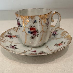 May include: A vintage teacup and saucer set. The white porcelain cup and saucer are decorated with floral patterns in red, blue, and pink, and edged with gold. The cup has a curved handle and a scalloped rim. The saucer has a matching design.