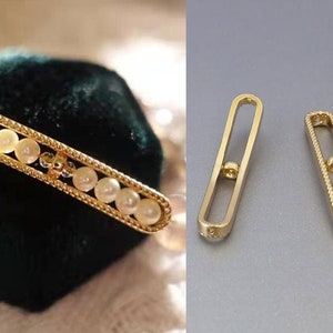 Könnte beinhalten: Goldfarbene Schmuckkomponenten. Ein Teil hat ein rechteckiges Design mit Perlen und einem dekorativen Rand. Die beiden anderen Teile sind längliche, offene Rahmenformen mit einem kleinen kreisförmigen Detail. Der Schmuck wird auf einer dunkelgrünen Oberfläche und einem grauen Hintergrund präsentiert.