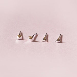 May include: Four pairs of small, silver-toned stud earrings with geometric designs. The earrings are arranged in a row on a light pink background. The designs include a heart, a triangle, and a square shape. The earrings are likely made of metal.