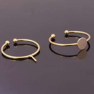 Può includere: Due braccialetti a polsino aperti color oro con un design minimalista. Un braccialetto ha un piccolo disco piatto e rotondo attaccato. Ogni braccialetto ha una piccola sfera su ogni estremità aperta. I braccialetti sono esposti su una superficie nera riflettente.