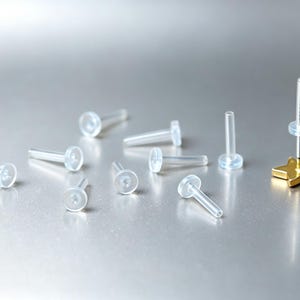 Peut inclure: Un ensemble de tiges de labret en plastique transparent et flexible avec des embouts plats circulaires, accompagné d'une tige en métal doré avec un motif d'étoile à cinq branches et un disque en silicone transparent.