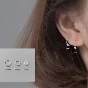 May include: Silver hoop earrings in various sizes are displayed. The image shows four different sizes of hoop earrings: 6mm, 8mm, 10mm, and 12mm. One pair of 10mm and 12mm earrings are worn in an ear. The earrings are simple and elegant.