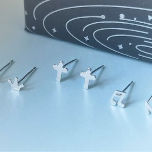 May include: Set of six silver stud earrings with different designs: a crown, a cross, a music note, and a heart. The earrings are on a light blue background.