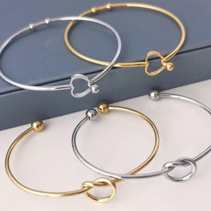 May include: Three silver and gold bangle bracelets with different charms. One bracelet has a heart charm, another has a knot charm, and the third has a simple ball charm.