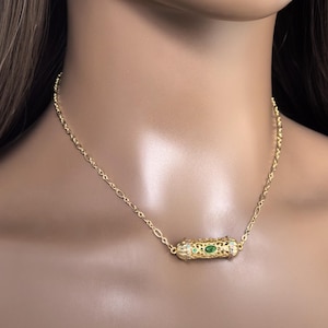 May include: A gold chain necklace with a small, ornate pendant featuring green gemstones. The pendant is shaped like a cylinder and has a filigree design.