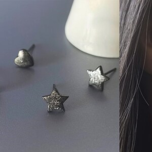 May include: Three pairs of silver stud earrings with a heart, star, and bar design. The earrings are shown on a gray surface and on a person's ear.