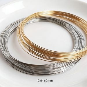 May include: Two spools of wire, one gold and one silver, are coiled on a white plate. The wire is 0.6mm x 60mm. The gold wire is a bright, shiny color, while the silver wire has a metallic sheen. The spools are side by side.