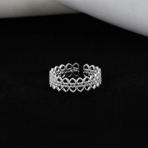 May include: A silver-colored adjustable ring with a delicate openwork design. The ring features a repeating pattern of heart-shaped cutouts, creating a lacy, elegant appearance. The ring is set against a black background.