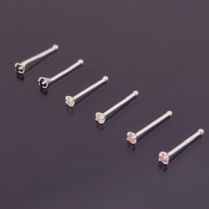 Può includere: Sei anelli per il naso in argento con piccole pietre preziose trasparenti e rosa. Gli anelli sono disposti in fila su una superficie grigio scuro.