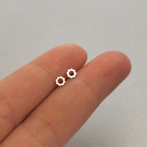 Pode incluir: Um par de brincos pequenos, dourados. Cada brinco apresenta um design de estrela aberta de oito pontas. Os brincos são exibidos na pele de uma pessoa, contra um fundo neutro. Joalharia.