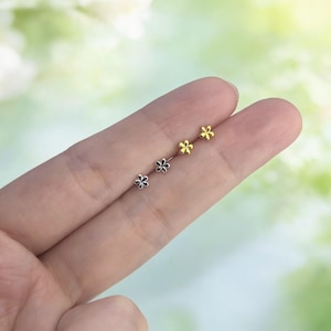 May include: Three pairs of flower-shaped stud earrings in silver and gold tones. The earrings are small and delicate, with a simple floral design. The earrings are displayed on a finger against a soft, blurred background.