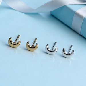 Puede incluir: Cuatro pendientes de botón con forma de media luna, dos dorados y dos plateados, se muestran sobre una superficie azul claro. Una caja de regalo azul claro con una cinta blanca está en el fondo. Los pendientes son pequeños y delicados.