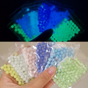 May include: Assortment of glow-in-the-dark beads in various colors, including green, blue, and white. The beads are packaged in clear plastic bags and are ready to be used for crafting projects.