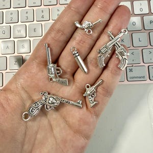 May include: A collection of silver-toned metal charms featuring various revolver designs, a single bullet-shaped charm, and a crossed-revolver charm. Each piece includes a small loop at the top for attachment, showcasing intricate detailing on the handles.
