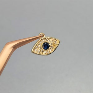 May include: Gold evil eye charm with a blue center and clear rhinestones around the eye. The charm is held by tweezers.