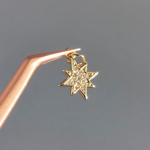 Op de afbeelding: Een gouden sterbedel met heldere zirkonia-stenen. De bedel wordt vastgehouden met een pincet.