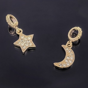 Op de afbeelding: Twee gouden bedels met heldere kristallen accenten. Een bedel is een stervorm en de andere is een halve maan.